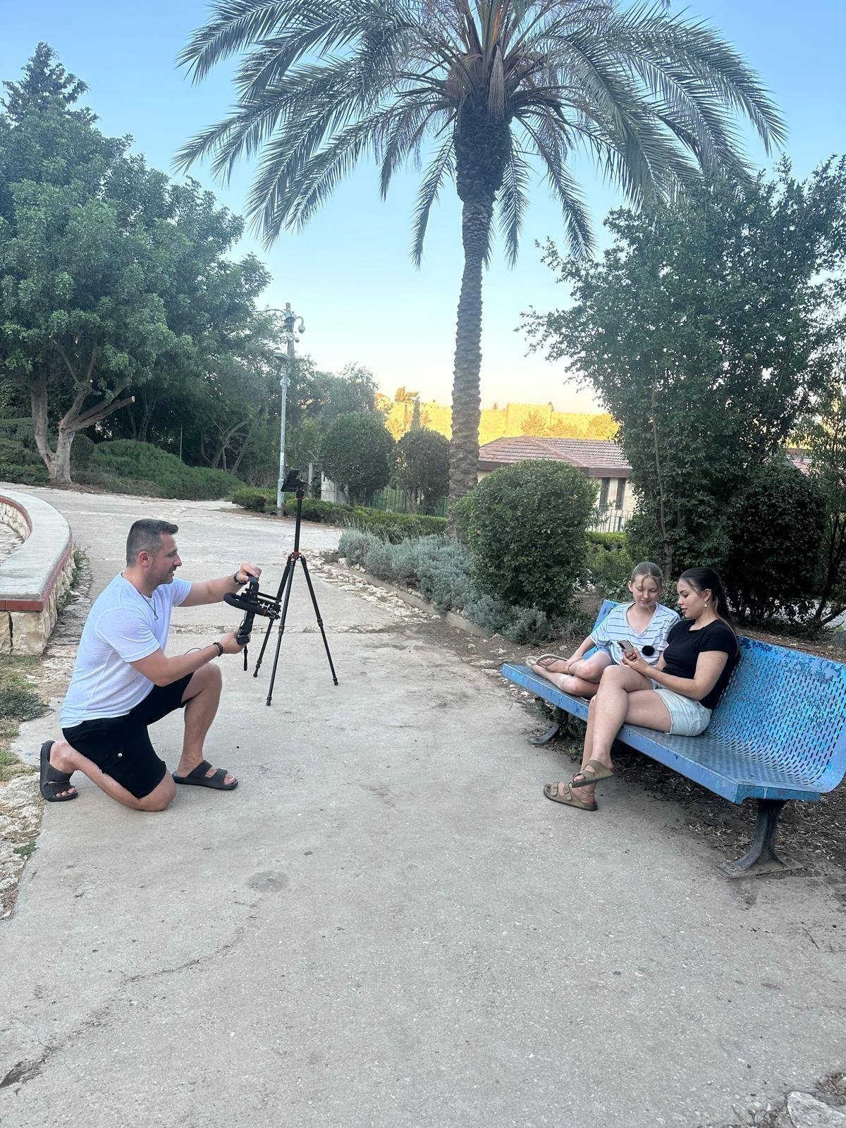 באמצע הפקה — צלם על חצובה מצלם שני אנשים שיושבים מול מצלמת וידאו בגן עצי דקל