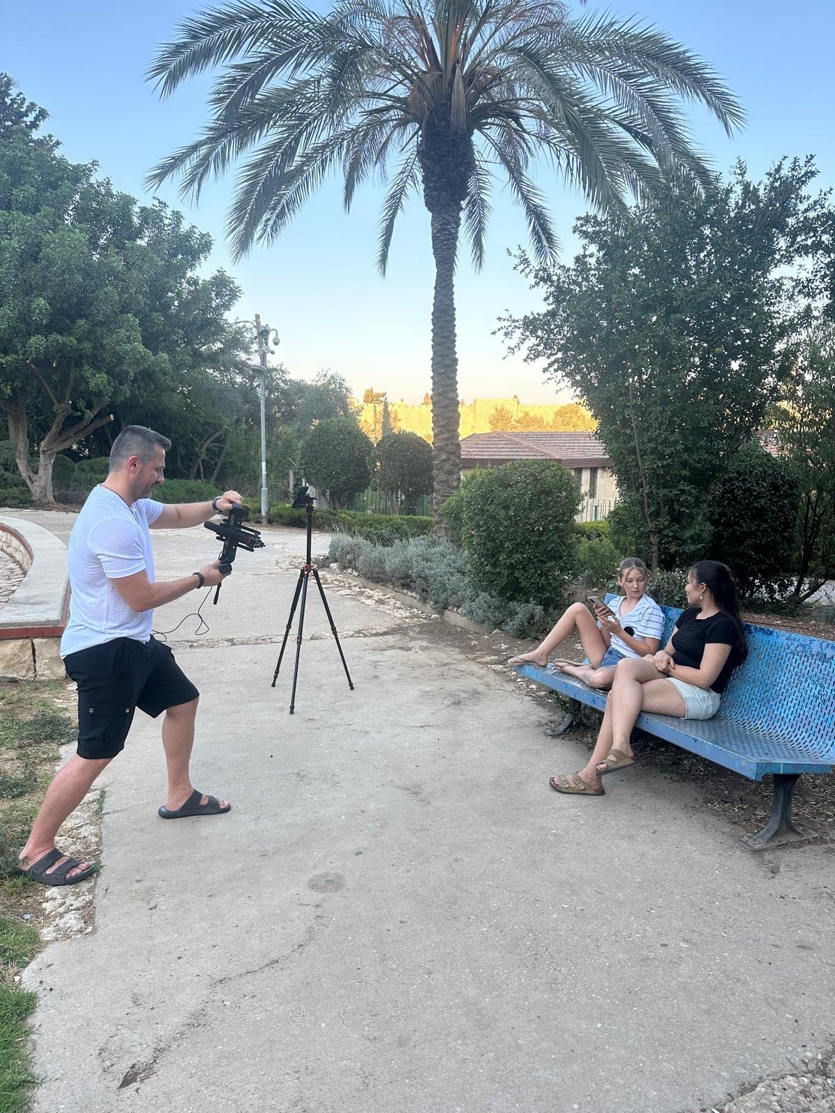 צלם מצלם שלושה אנשים שיושבים על ספסל בפארק עם עץ דקל ברקע