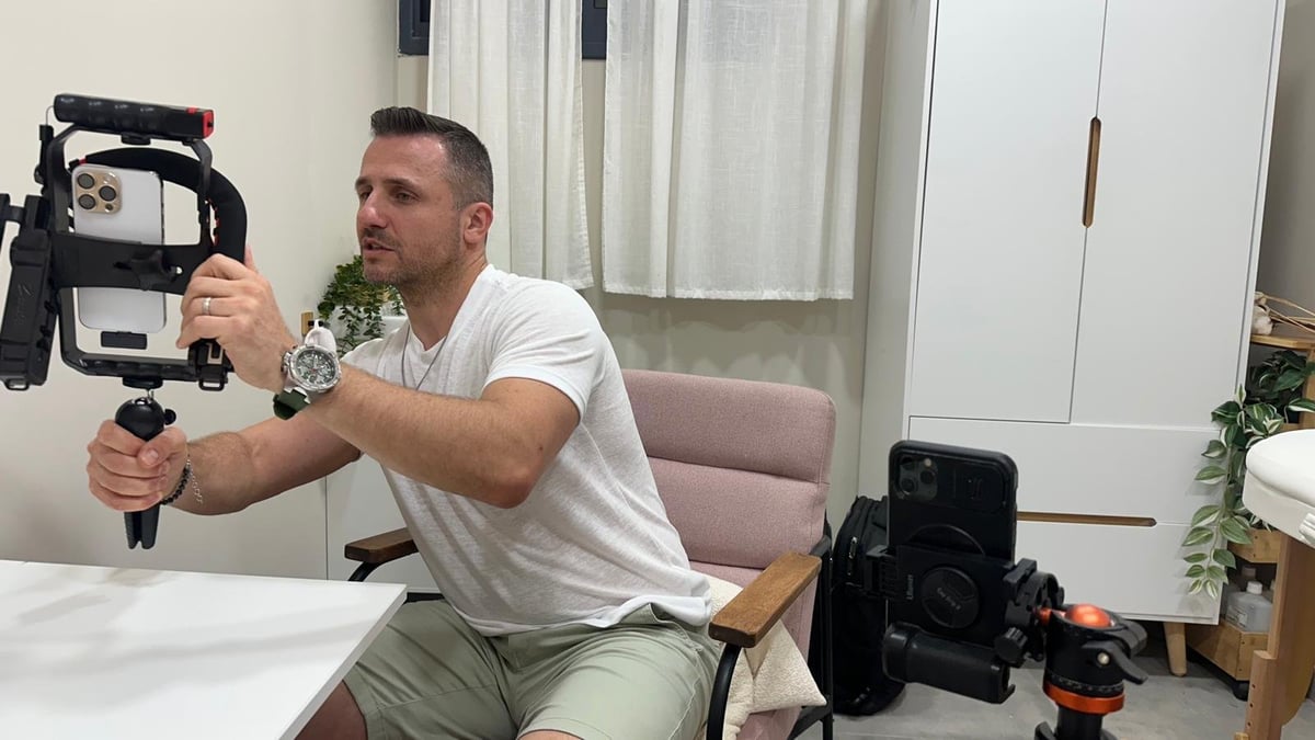 איש בחולצה לבנה מחזיק מייצב מצלמה מקצועי בסטודיו ביתי עם ציוד על השולחן